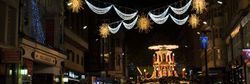 A festive street in Birmingham adorned with glowing Christmas lights, including golden snowflakes and draped garlands, with a vibrant holiday market in the background buzzing with activity.