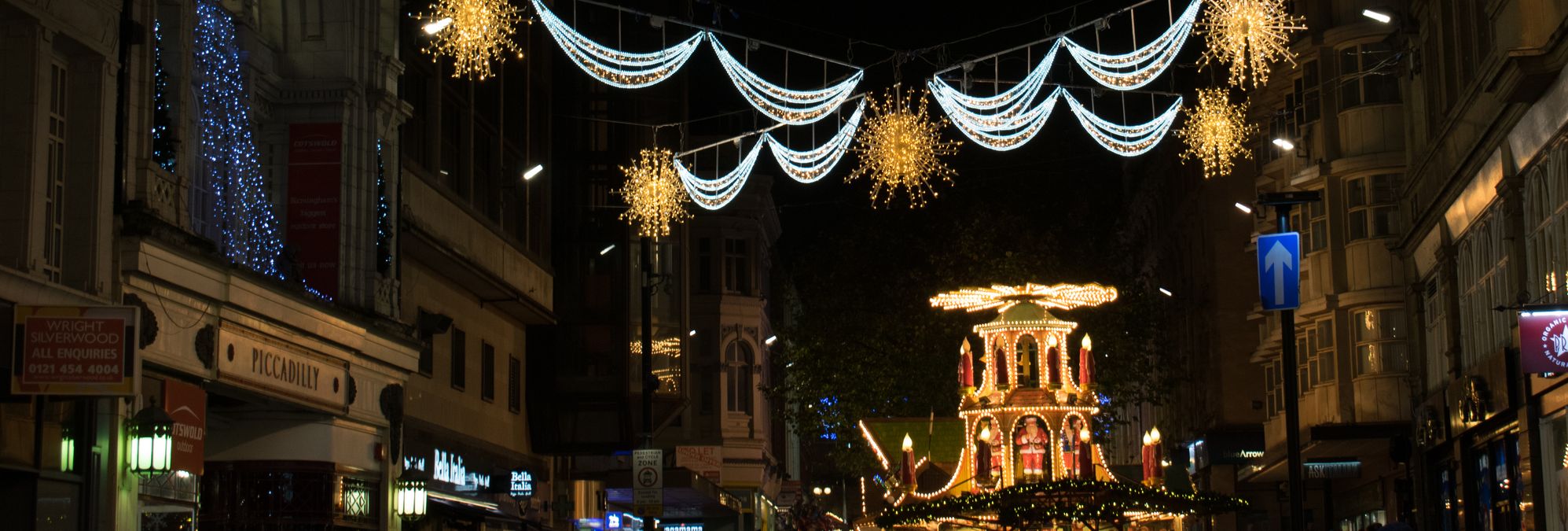 A festive street in Birmingham adorned with glowing Christmas lights, including golden snowflakes and draped garlands, with a vibrant holiday market in the background buzzing with activity.