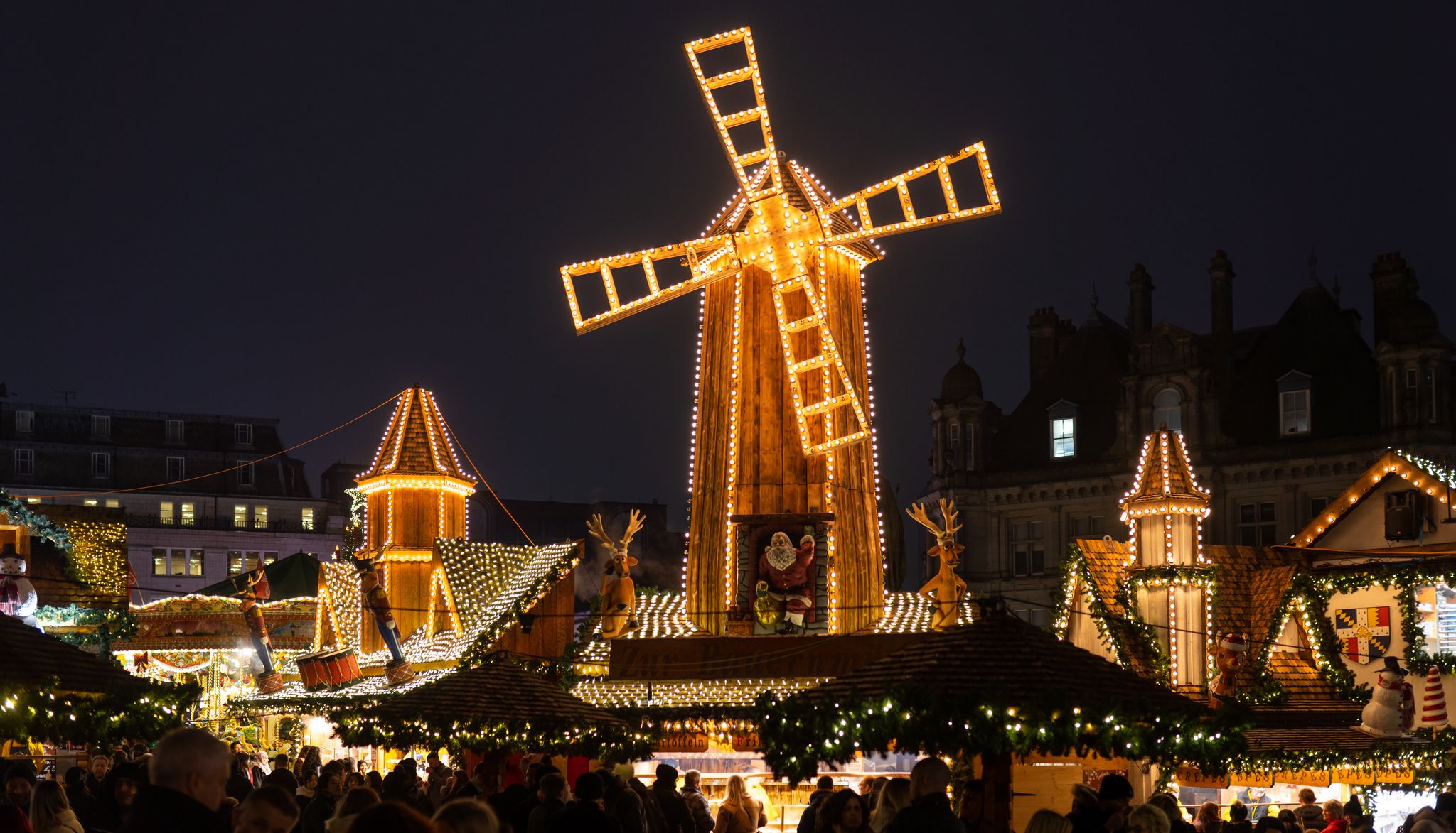 A festive Christmas market in Birmingham at night, featuring a brightly lit wooden windmill, decorated stalls, and holiday ornaments, bustling with visitors enjoying the seasonal cheer.