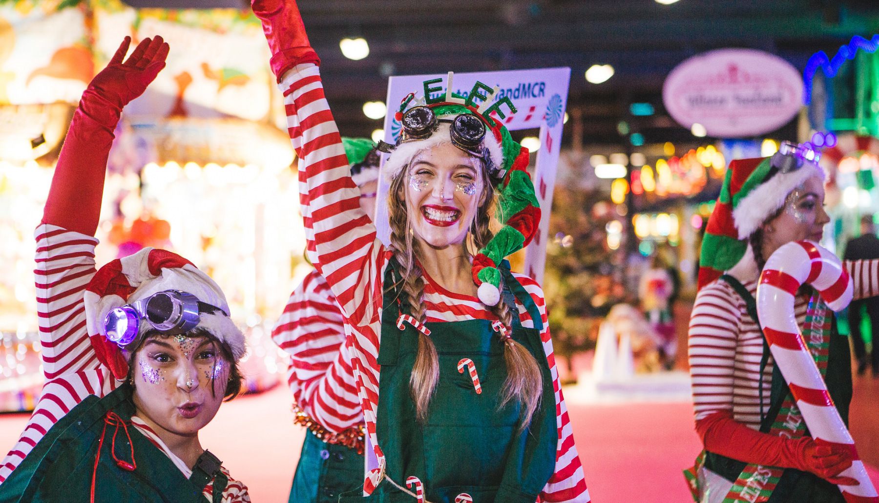 A group of cheerful elves in festive outfits with candy canes and glittery faces, celebrating Christmas in Birmingham, set against a colorful backdrop of holiday lights and decorations.
