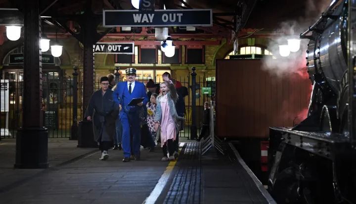 Children and a conductor in a blue uniform, walk along a festive, vintage train platform with steam rising from a locomotive, capturing the magic of Christmas in Birmingham.