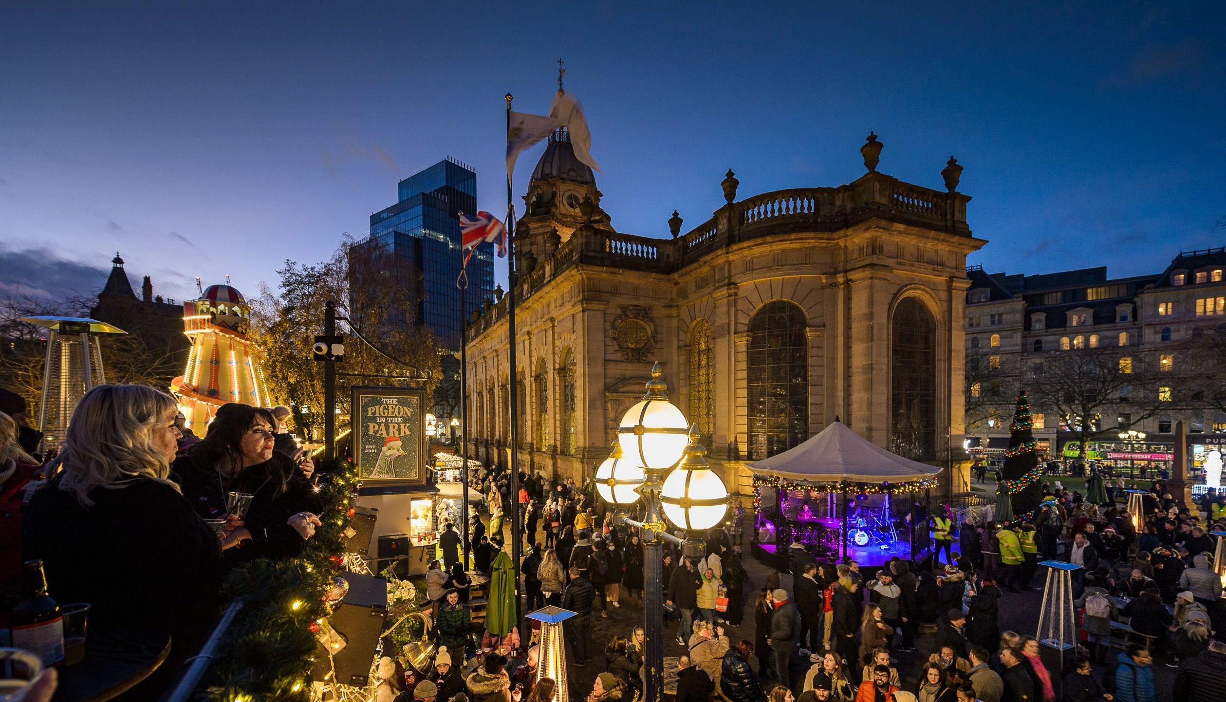A vibrant Christmas market in Birmingham, set around a historic cathedral, features festive lights, a towering helter-skelter, and bustling crowds enjoying the holiday spirit and live music.