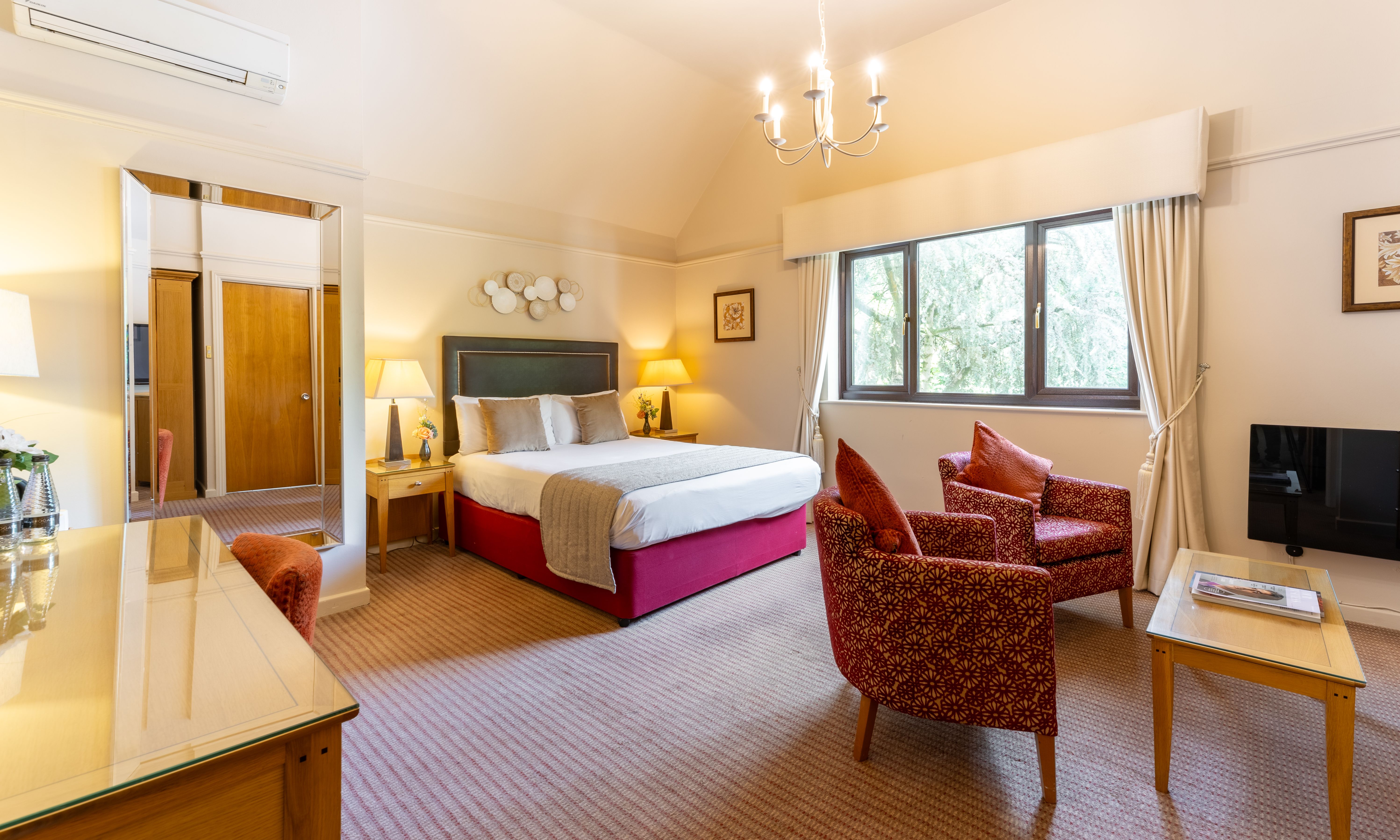 Spacious bedroom with a large bed, two patterned armchairs, a desk, a mirror, bedside tables, and a flat-screen TV.