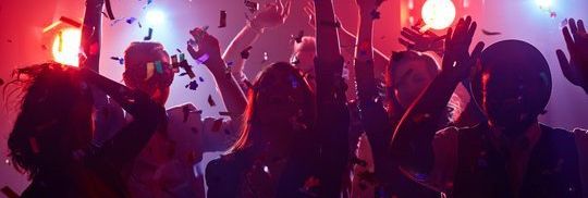 People dancing with raised hands under colourful lights as confetti falls at a party