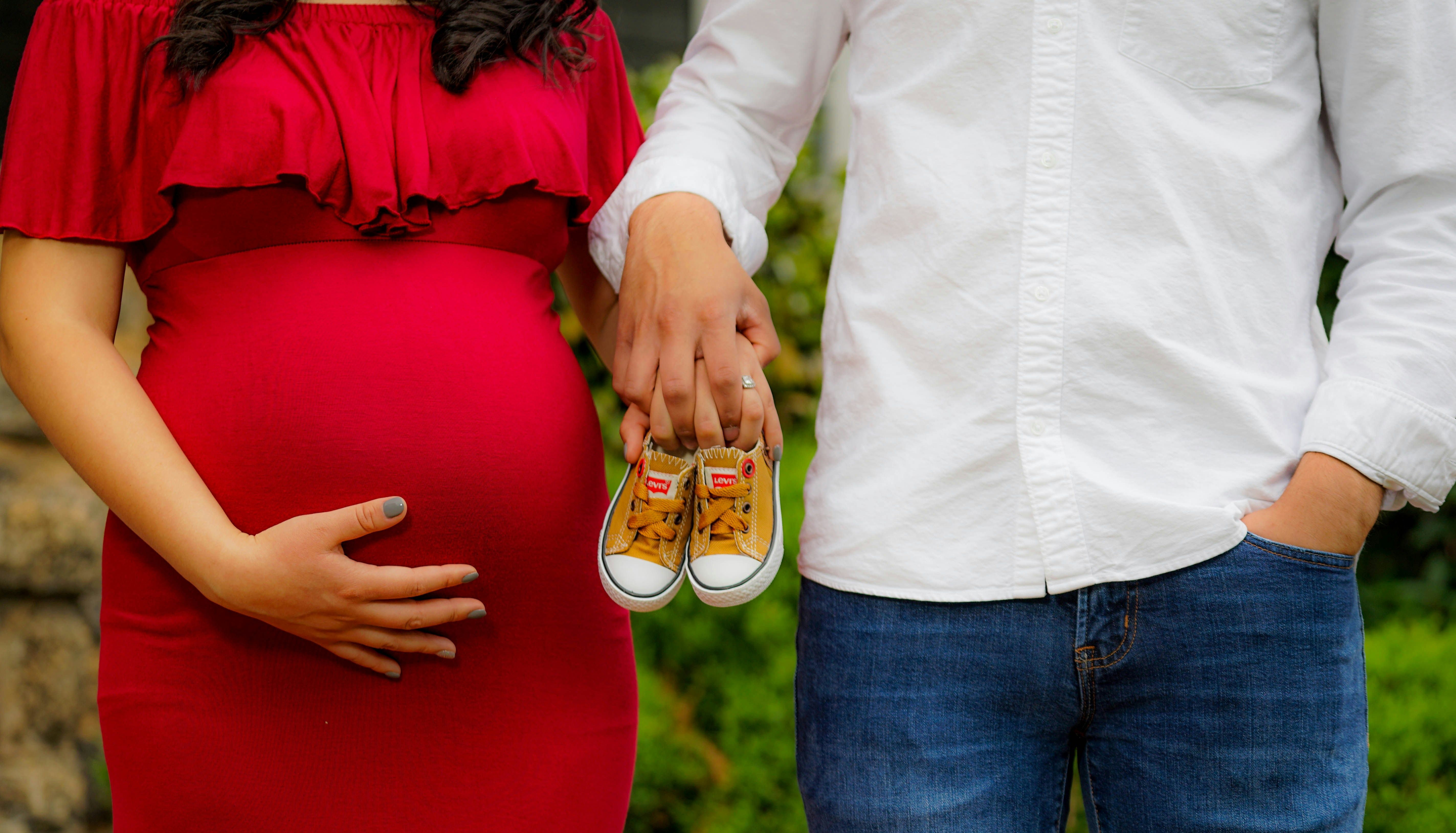 Pregnant woman in a red dress with partner holding baby shoes