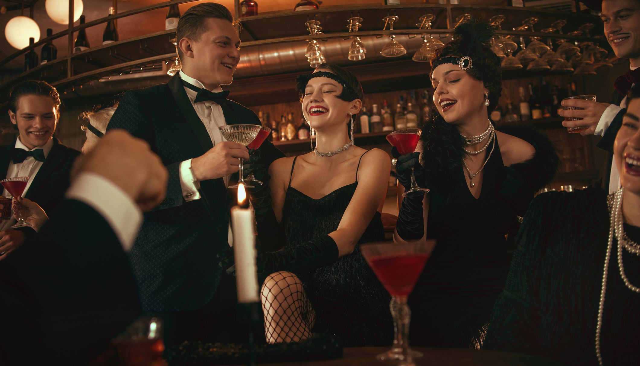 Friends in 1920s-inspired eveningwear laughing and toasting with cocktails in a warmly lit bar.