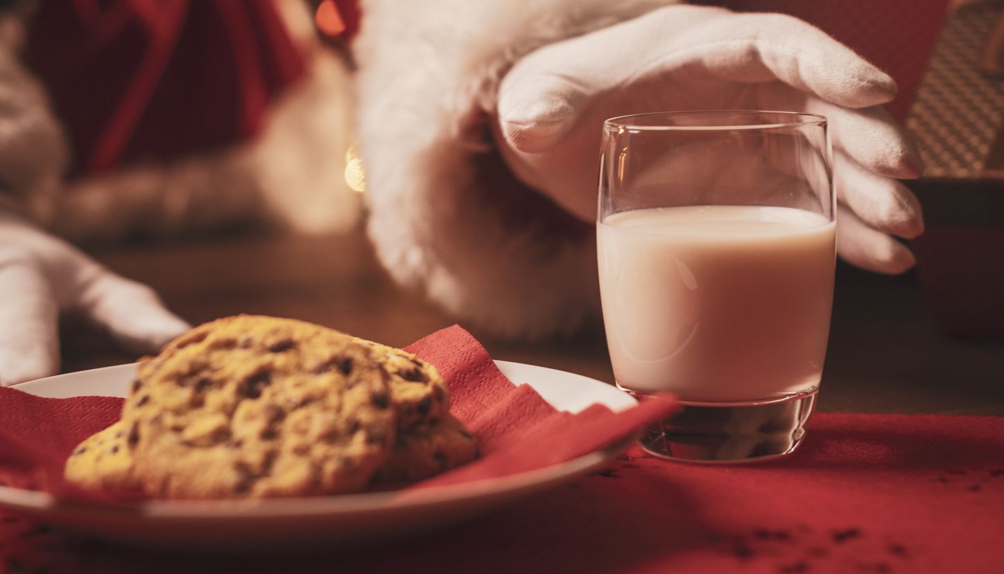 Santa in red suit reaching for a glass of milk beside a plate of biscuits, with a Christmas tree blurred in the background