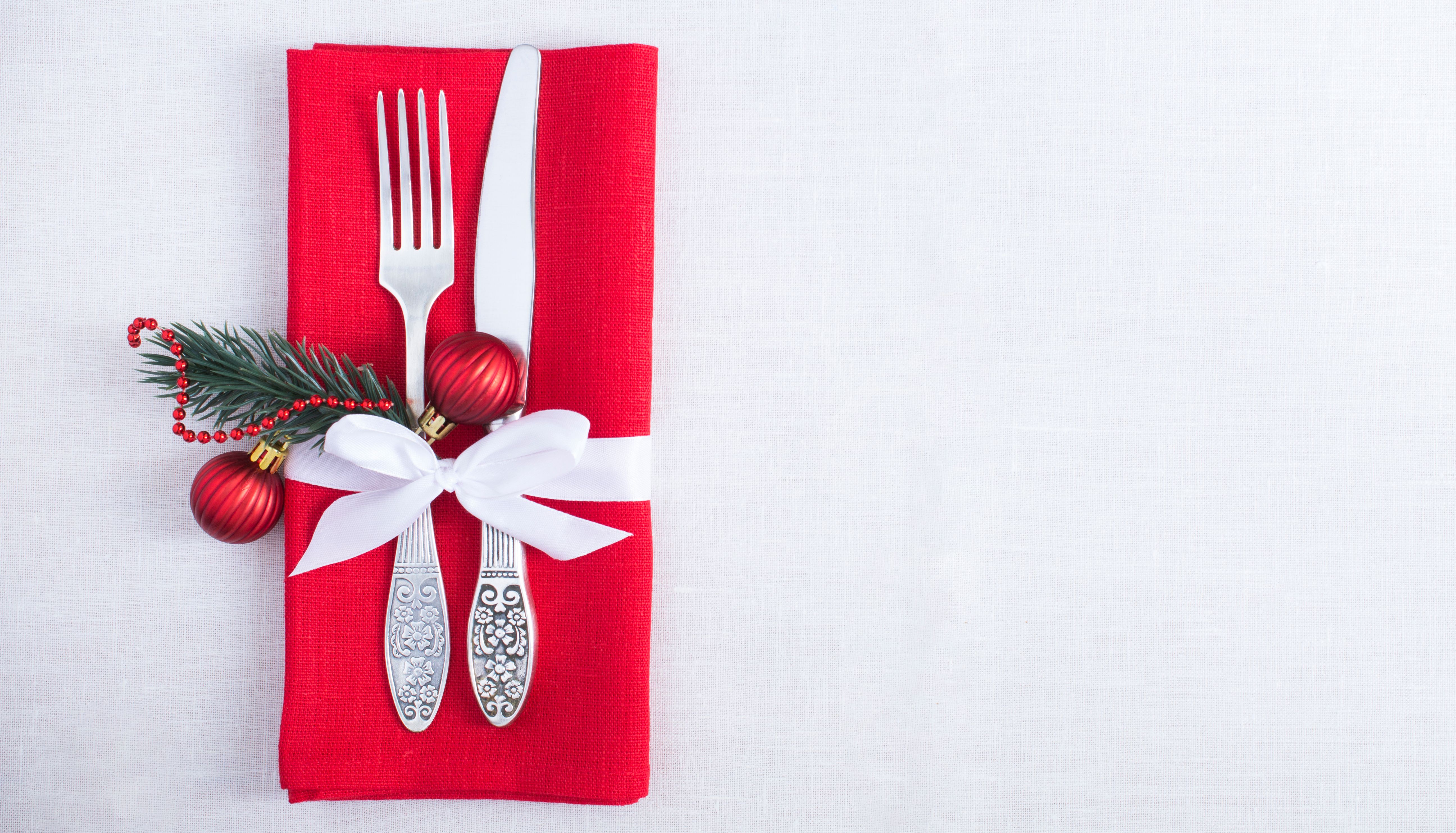 Christmas table setting with fork and knife on a red napkin tied with a white ribbon, decorated with fir sprig, red baubles and bead garland on a white tablecloth