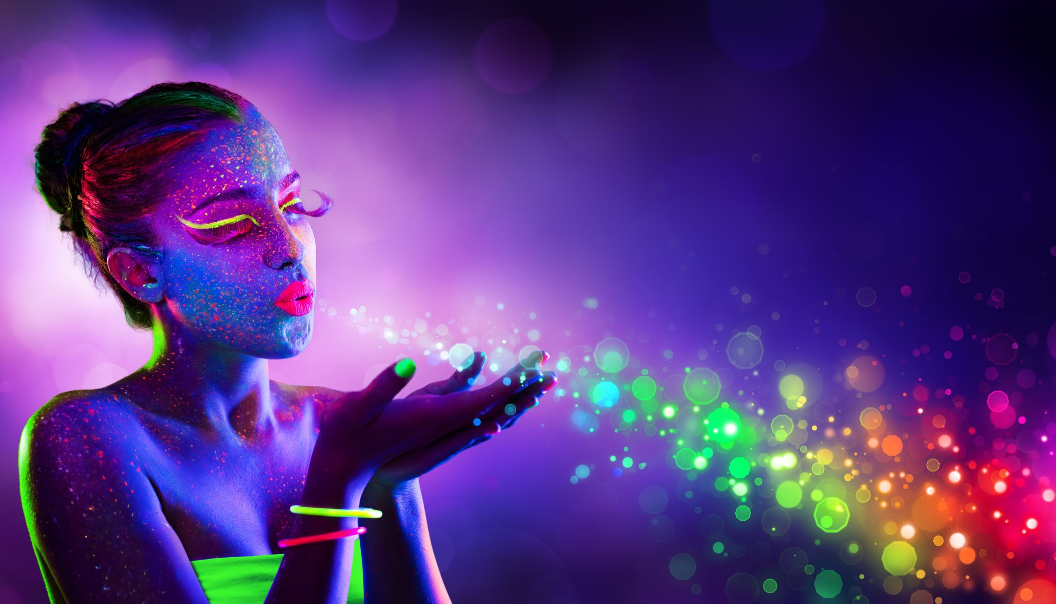 Woman with neon UV body paint blowing sparkling rainbow light from her hands against a purple background
