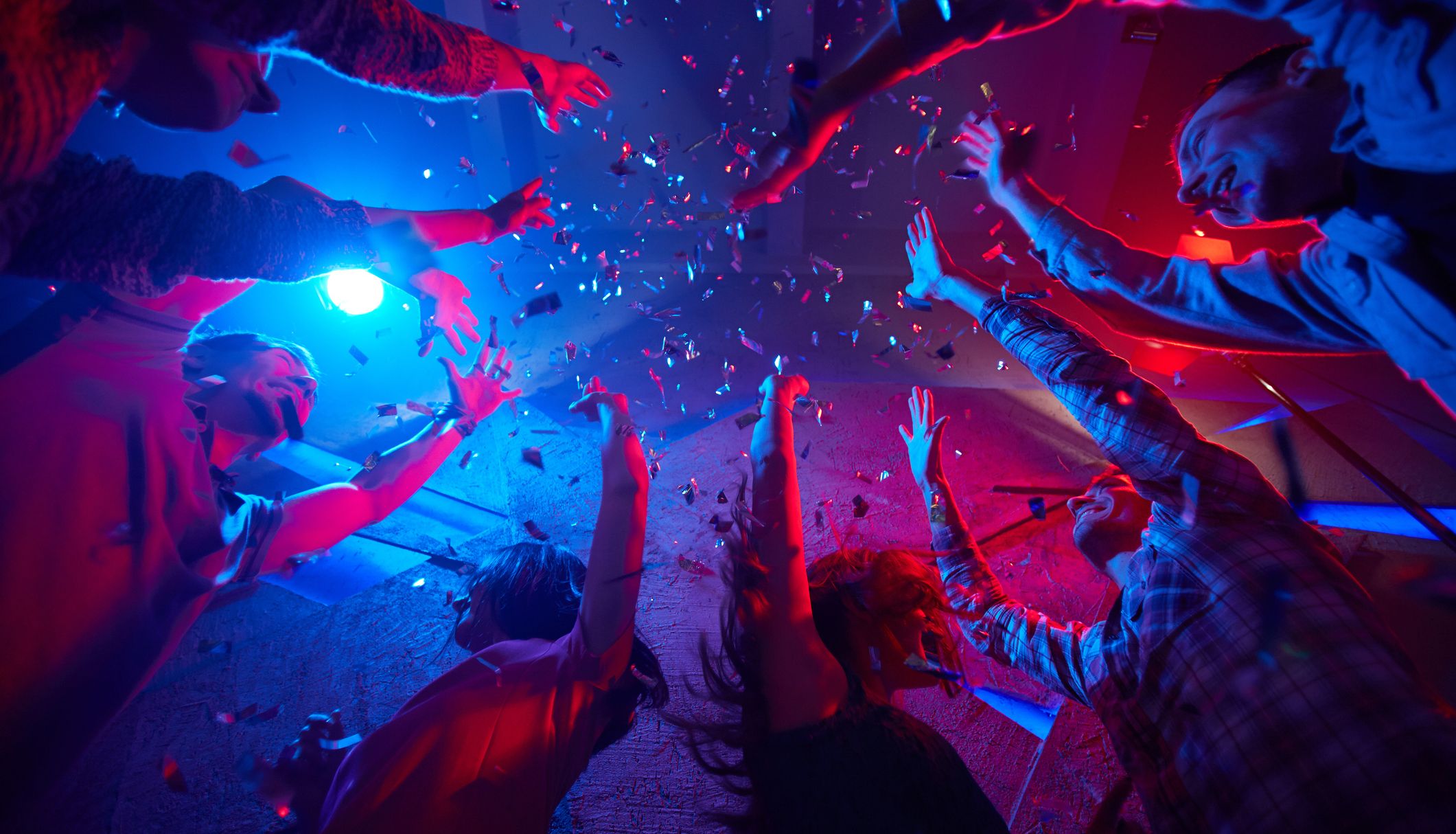 Group of friends reaching up towards falling confetti under blue and red party lights