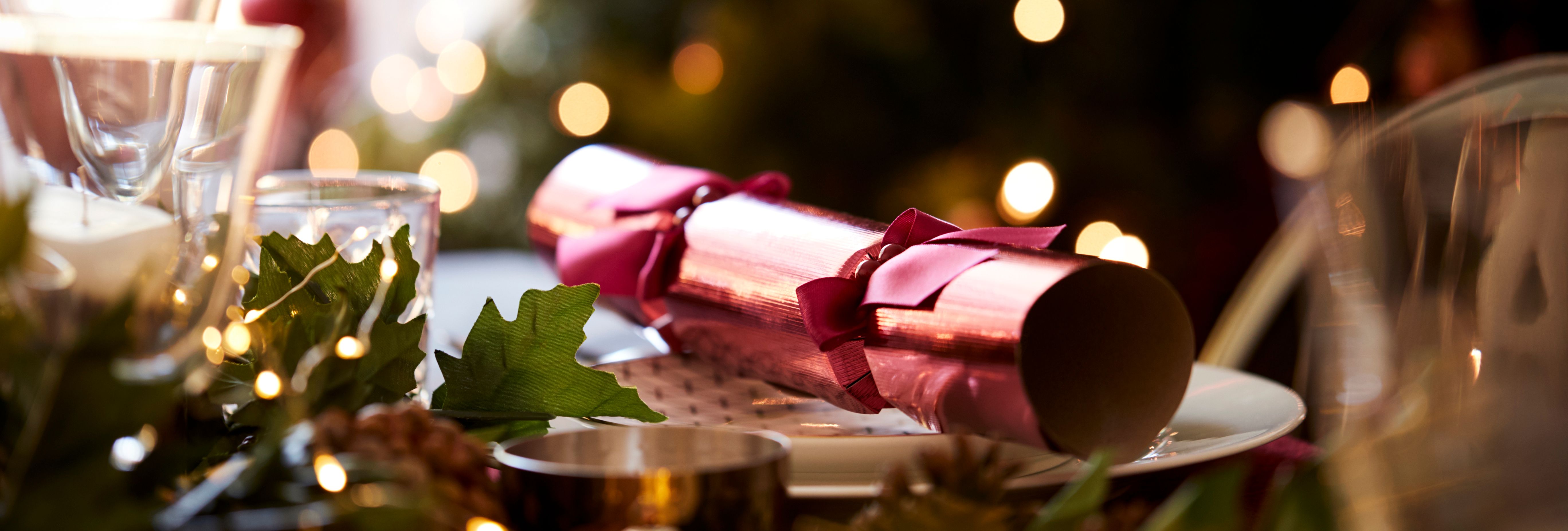 Christmas table setting with pink cracker, festive greenery and blurred fairy lights in the background