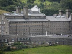 A grey stone prison with barred windows and chimneys stands behind suburban homes, set against a backdrop of green hills and dense trees.