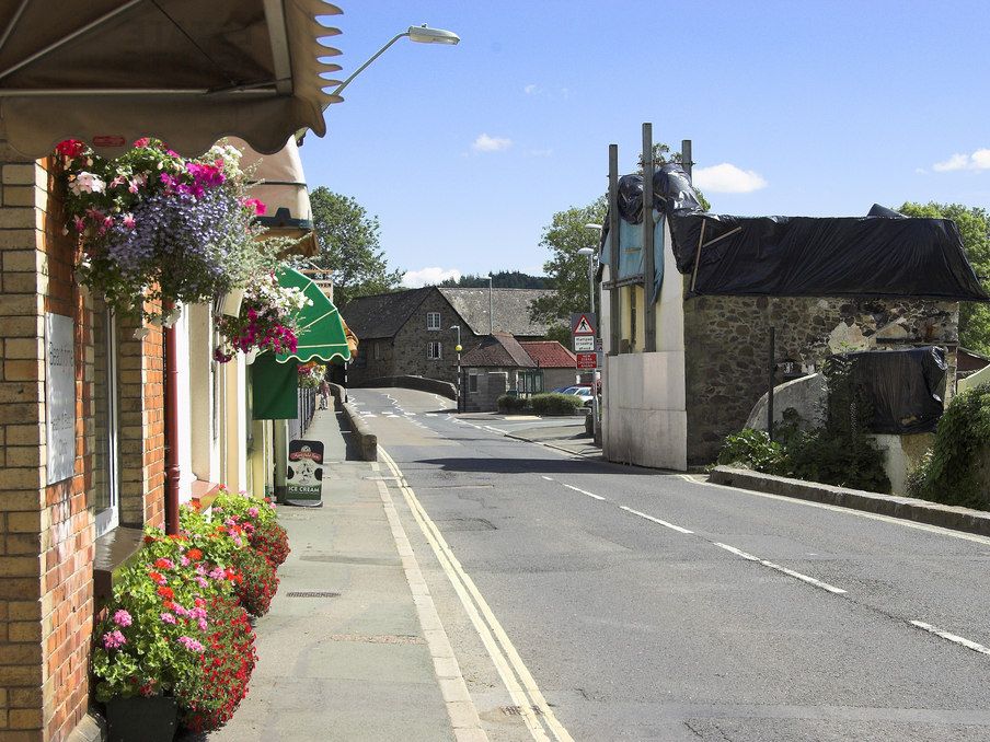 A sunny street with hanging flower baskets and shop awnings on the left, and a partially ruined stone building covered with tarps on the right.
