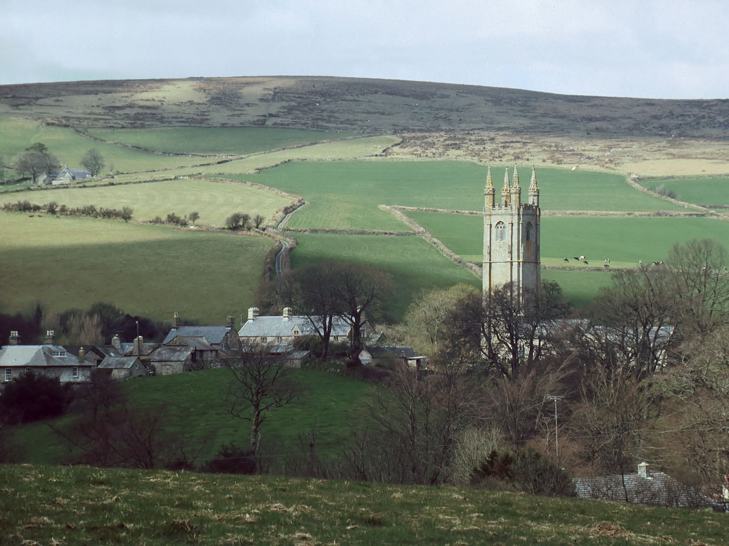 A stone church tower rises above trees and cottages in a rural valley, surrounded by rolling green fields and hills under a cloudy sky.