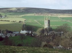 A stone church tower rises above trees and cottages in a rural valley, surrounded by rolling green fields and hills under a cloudy sky.