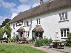 A white thatched-roof pub with picnic tables and parasols in a grassy garden, set against a blue sky with scattered clouds on a sunny day.