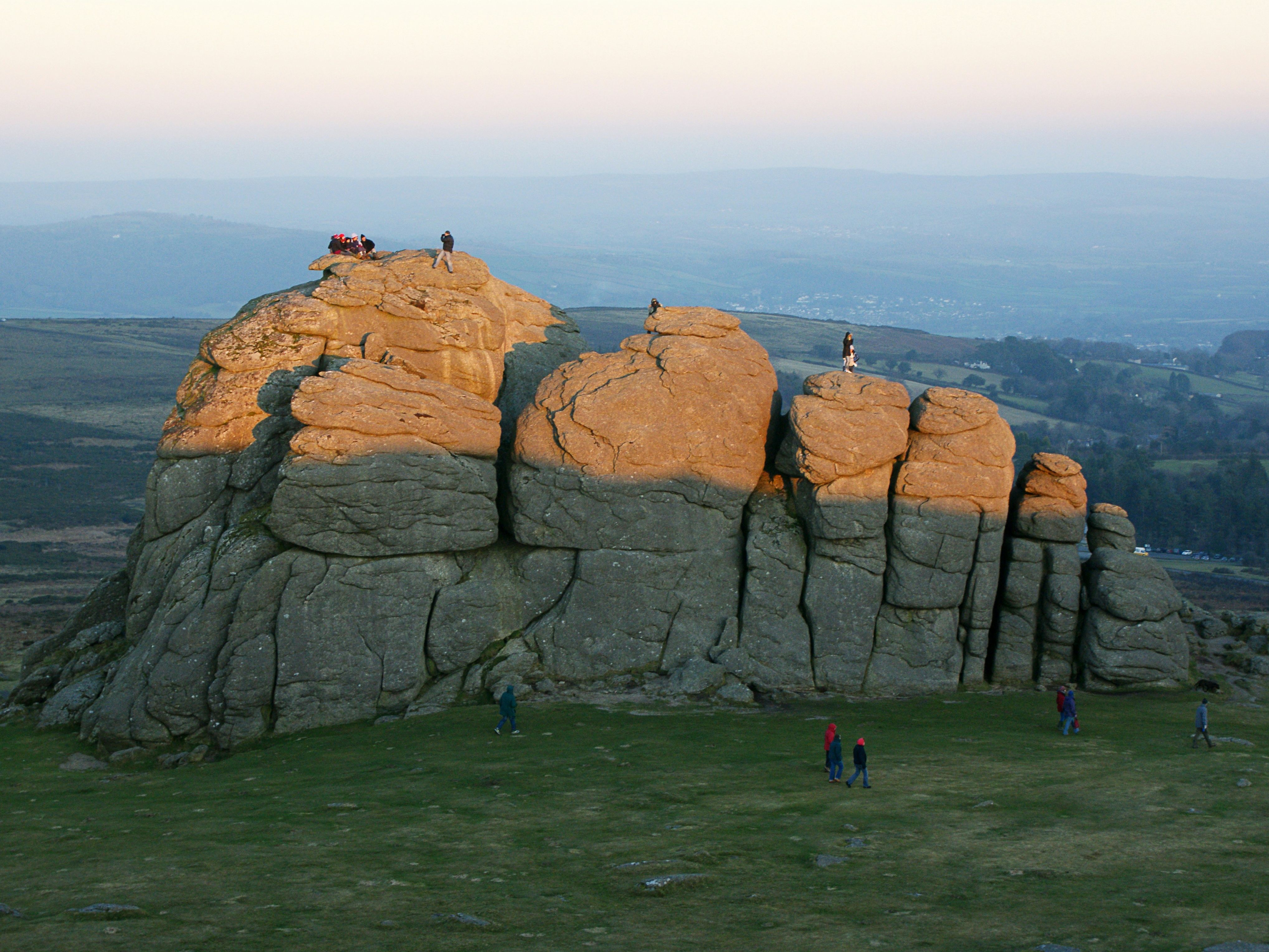 People climb and explore Haytor, a large granite rock formation on Dartmoor, lit by golden sunset light with rolling hills in the background.