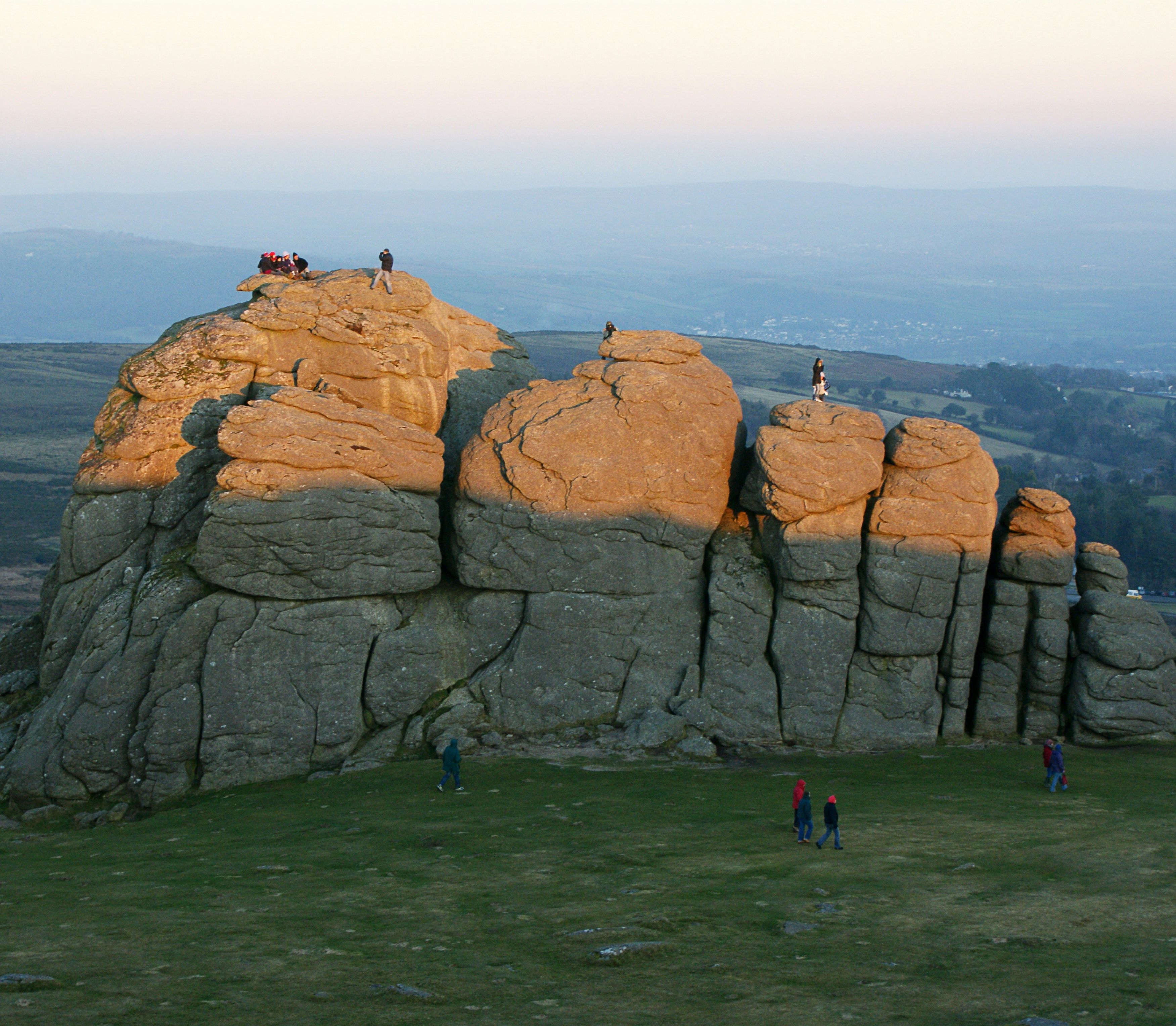 People climb and explore Haytor, a large granite rock formation on Dartmoor, lit by golden sunset light with rolling hills in the background.