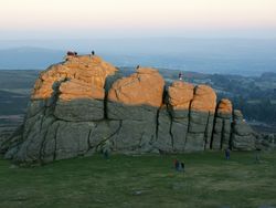People climb and explore Haytor, a large granite rock formation on Dartmoor, lit by golden sunset light with rolling hills in the background.