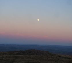 A twilight view of a moonlit sky over rolling hills, with a large ancient stone cairn in the foreground and soft pink hues along the horizon.