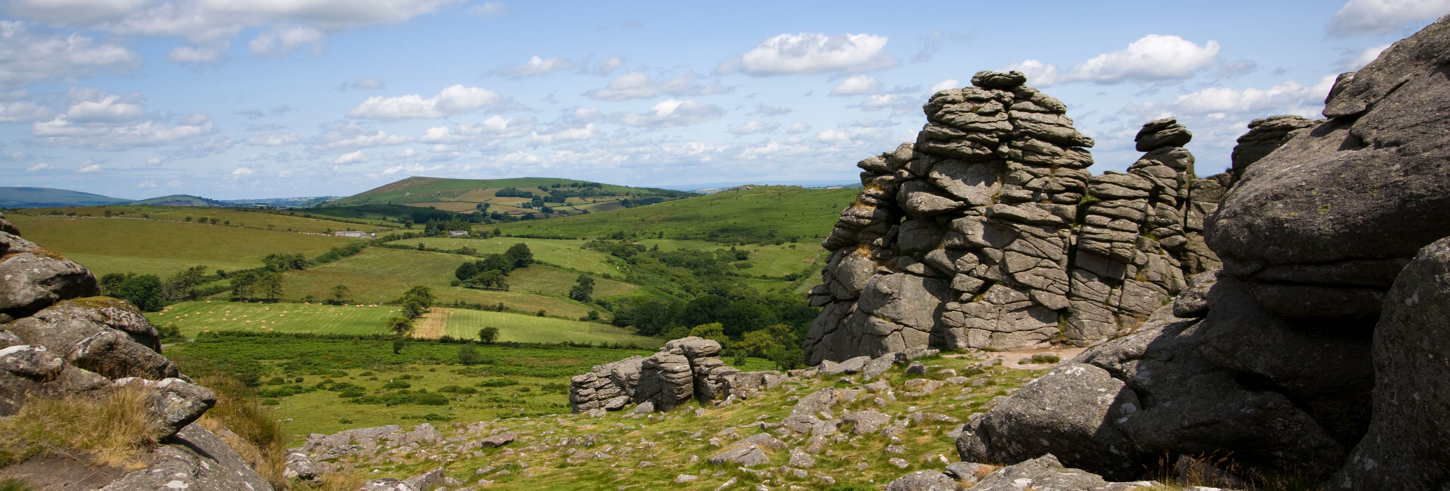 Rocky tors rise above a lush green Dartmoor landscape under a blue sky with scattered clouds, offering a scenic view of the rolling countryside.