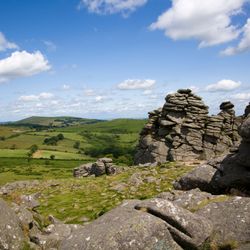 Rocky tors rise above a lush green Dartmoor landscape under a blue sky with scattered clouds, offering a scenic view of the rolling countryside.