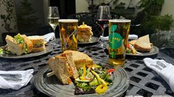 A selection of sandwiches, salads, wine, and cider served on a sunny outdoor table, perfect for a relaxed al fresco lunch in a garden setting.