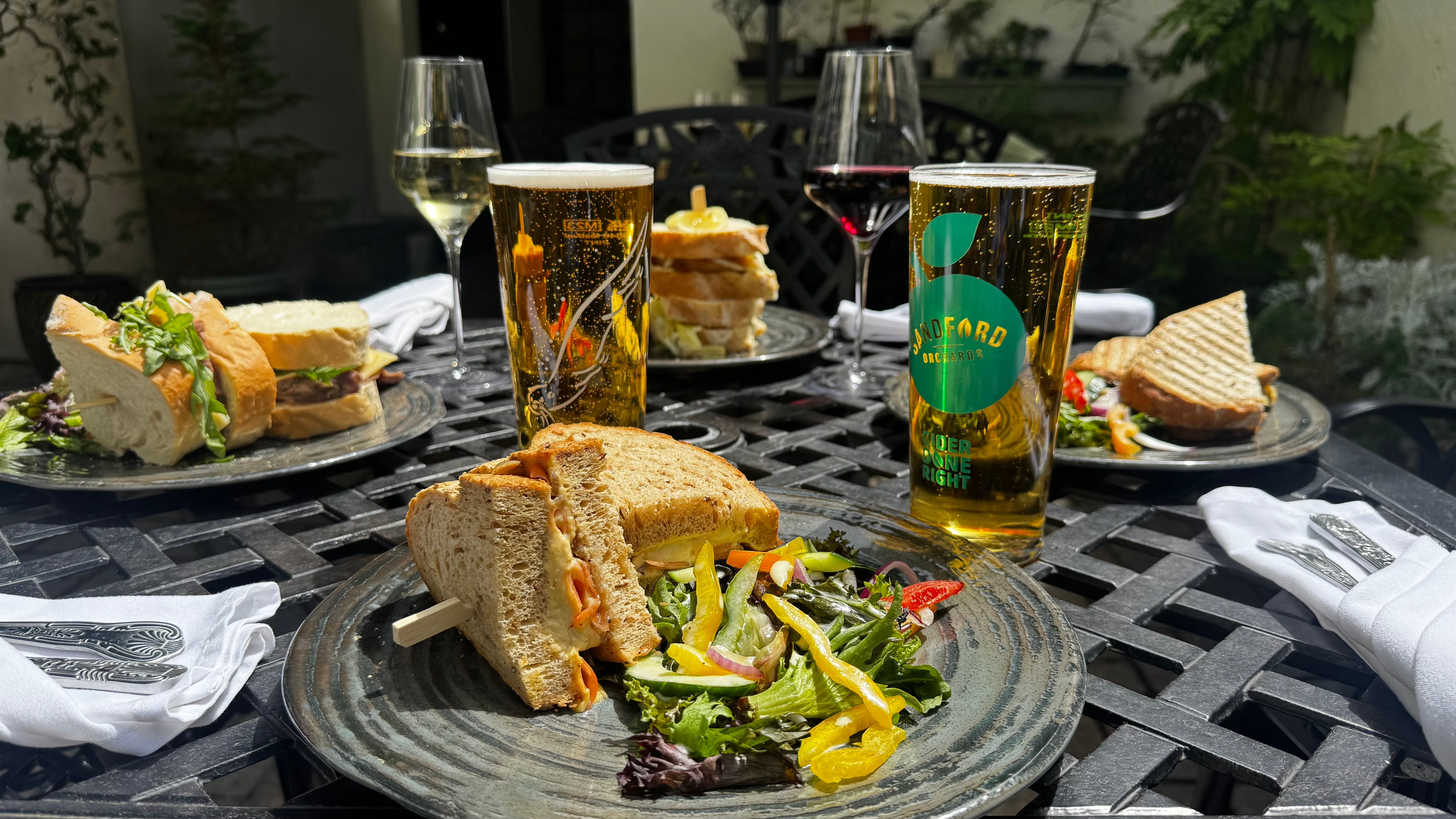 A selection of sandwiches, salads, wine, and cider served on a sunny outdoor table, perfect for a relaxed al fresco lunch in a garden setting.