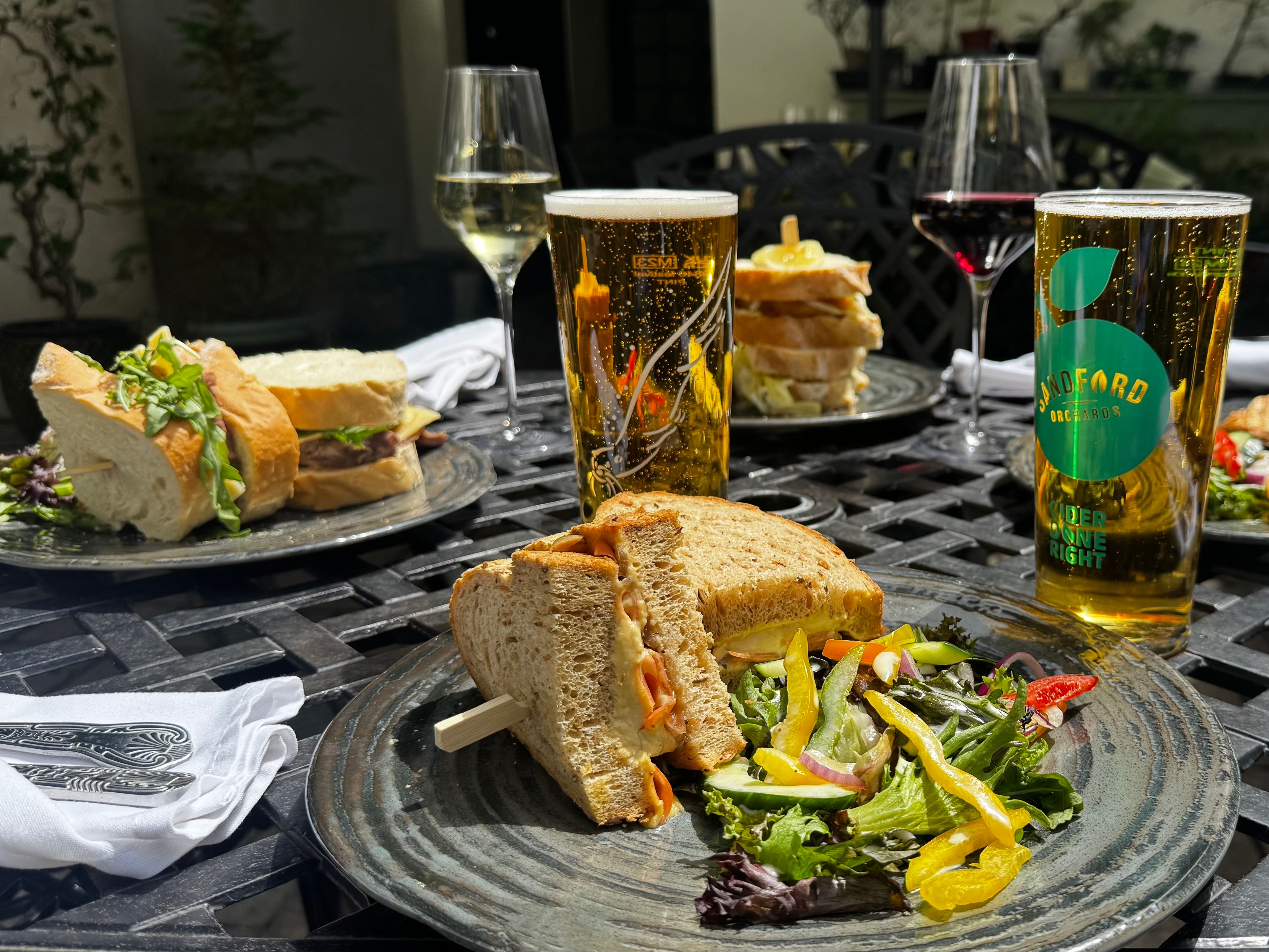 A selection of sandwiches, salads, wine, and cider served on a sunny outdoor table, perfect for a relaxed al fresco lunch in a garden setting.