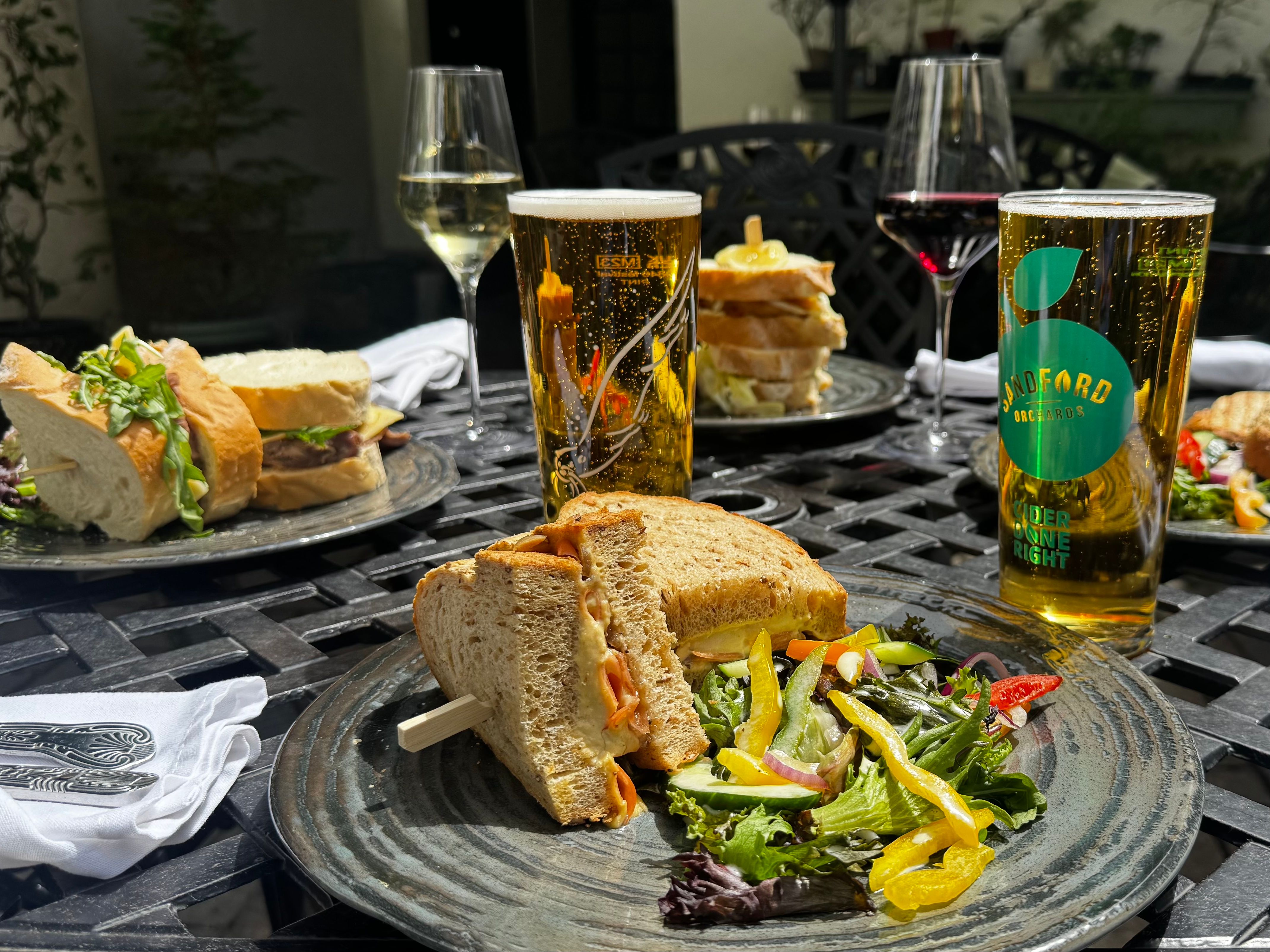 A selection of sandwiches, salads, wine, and cider served on a sunny outdoor table, perfect for a relaxed al fresco lunch in a garden setting.