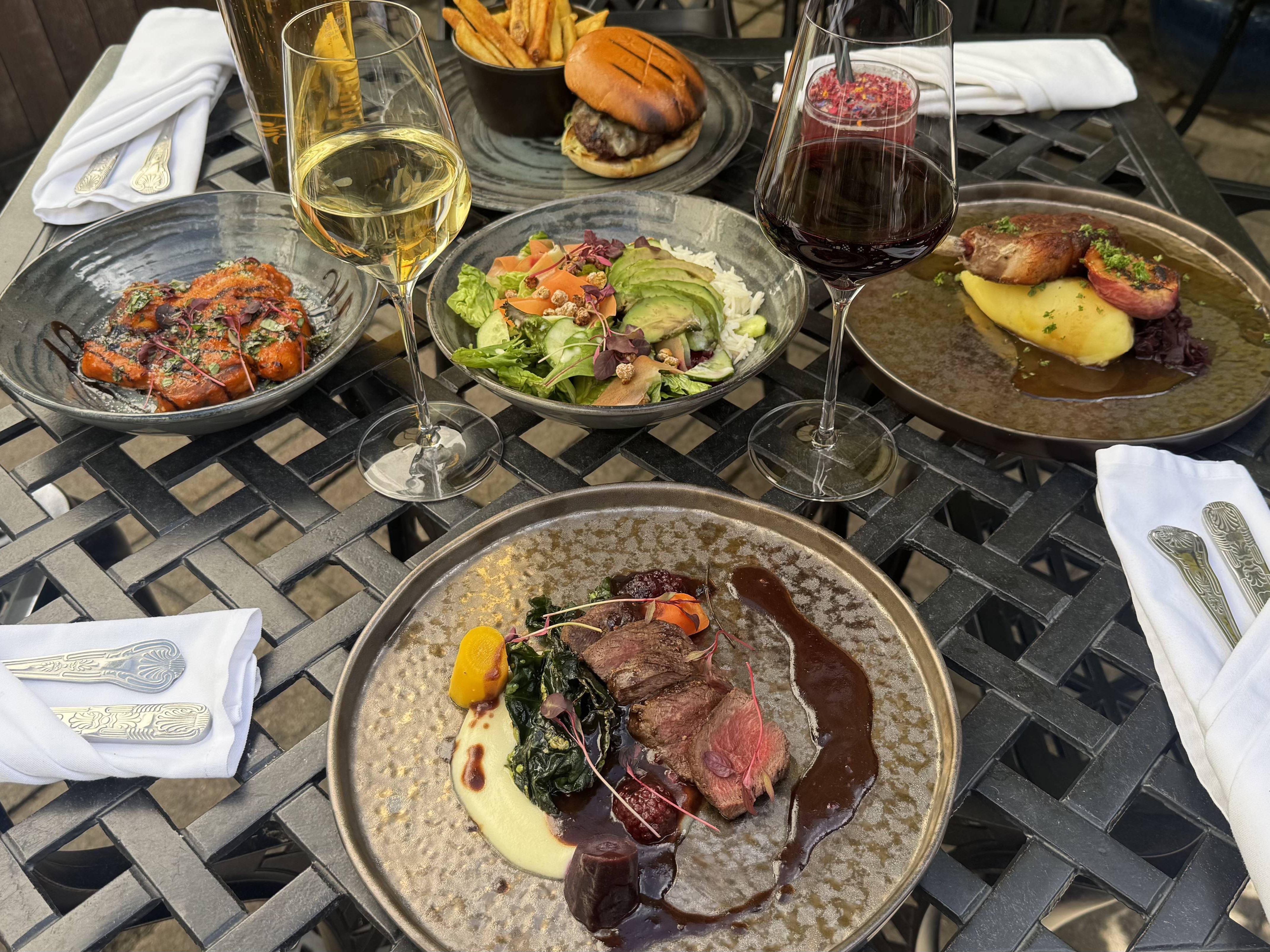 A gourmet outdoor meal with wine, showcasing elegant mains, starters, and sides on a wrought iron table, ready for al fresco dining in the sun.