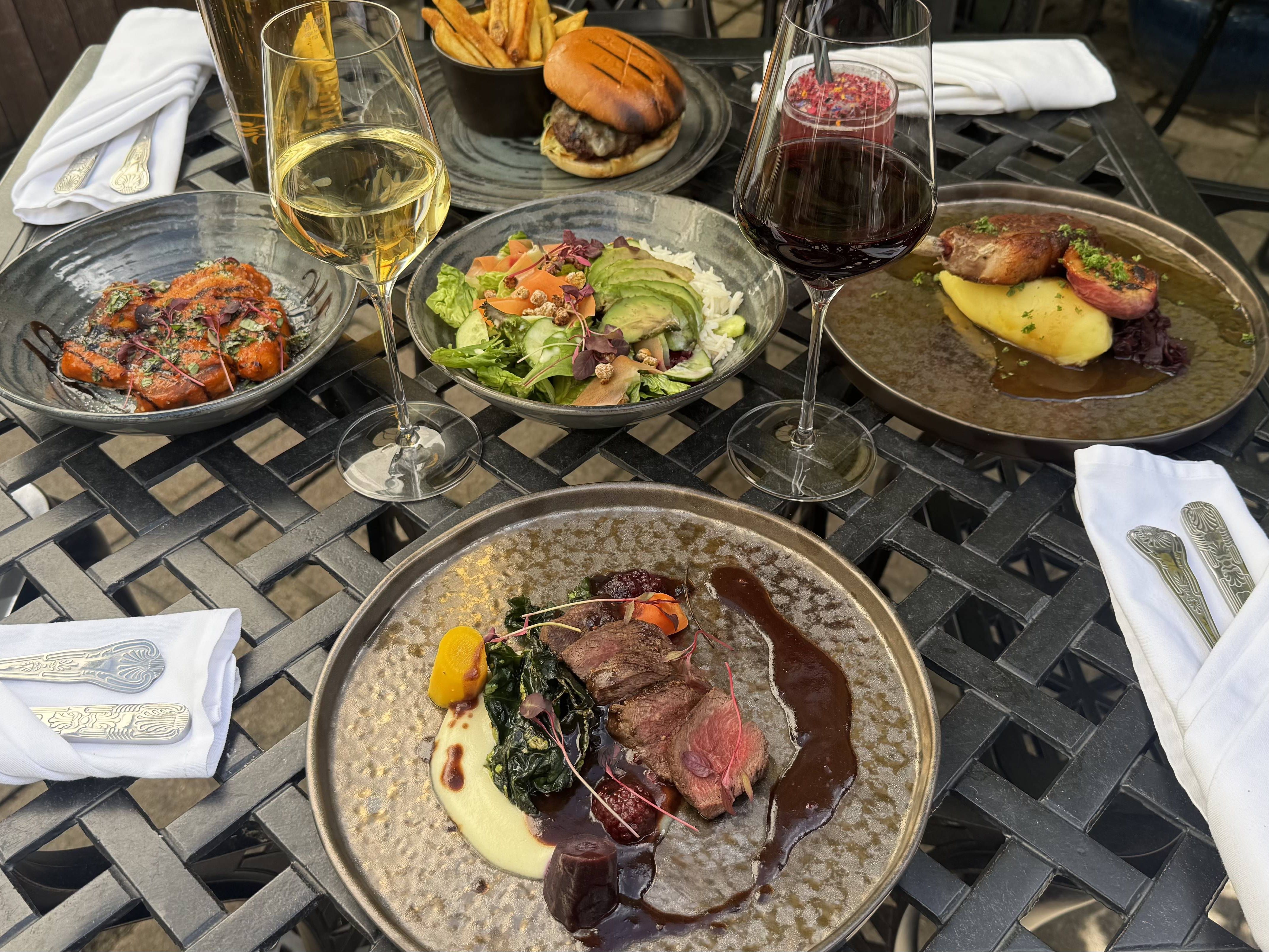 A gourmet outdoor meal with wine, showcasing elegant mains, starters, and sides on a wrought iron table, ready for al fresco dining in the sun.