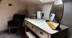 A modern dressing table setup with a mirror, kettle, cups, and a dark green velvet chair with a pillow.