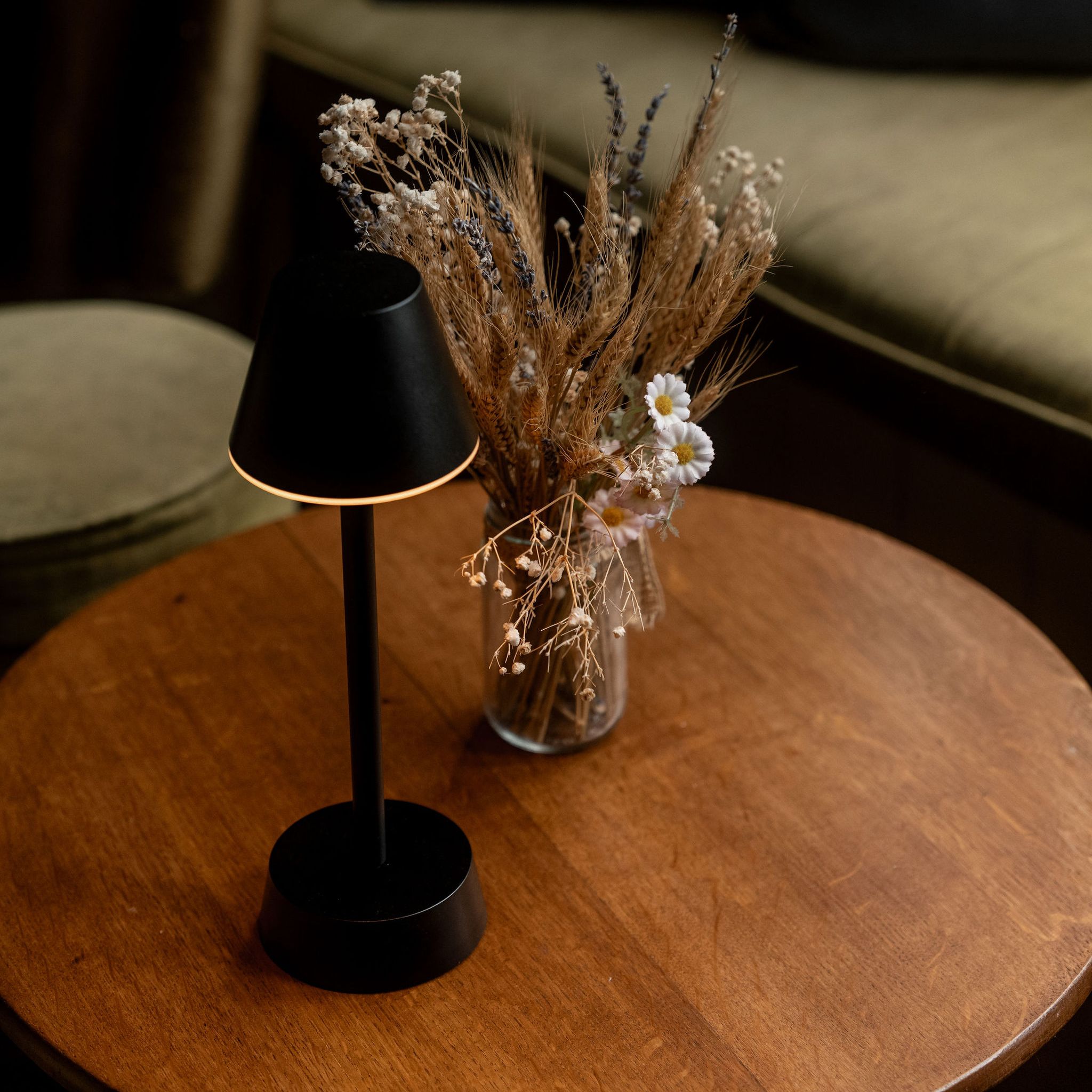 A cozy table with a black lamp and a vase of dried flowers, next to a cushioned seating area.