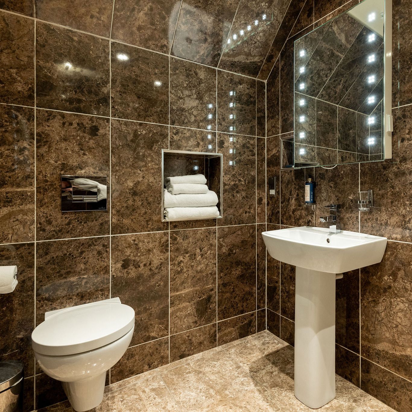 Modern bathroom with brown marble walls and floor, wall-mounted toilet, pedestal sink, illuminated mirror, niche with neatly stacked white towels, and chrome fixtures.