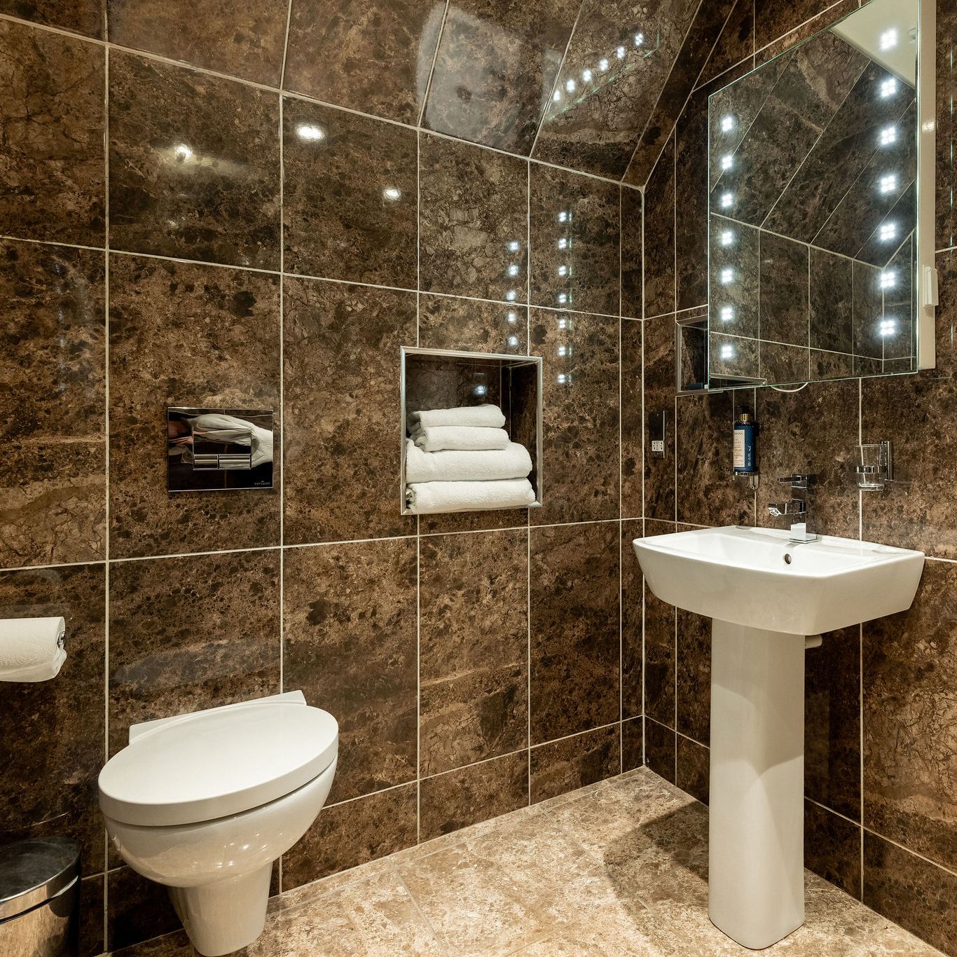 Modern bathroom with brown marble walls and floor, wall-mounted toilet, pedestal sink, illuminated mirror, niche with neatly stacked white towels, and chrome fixtures.