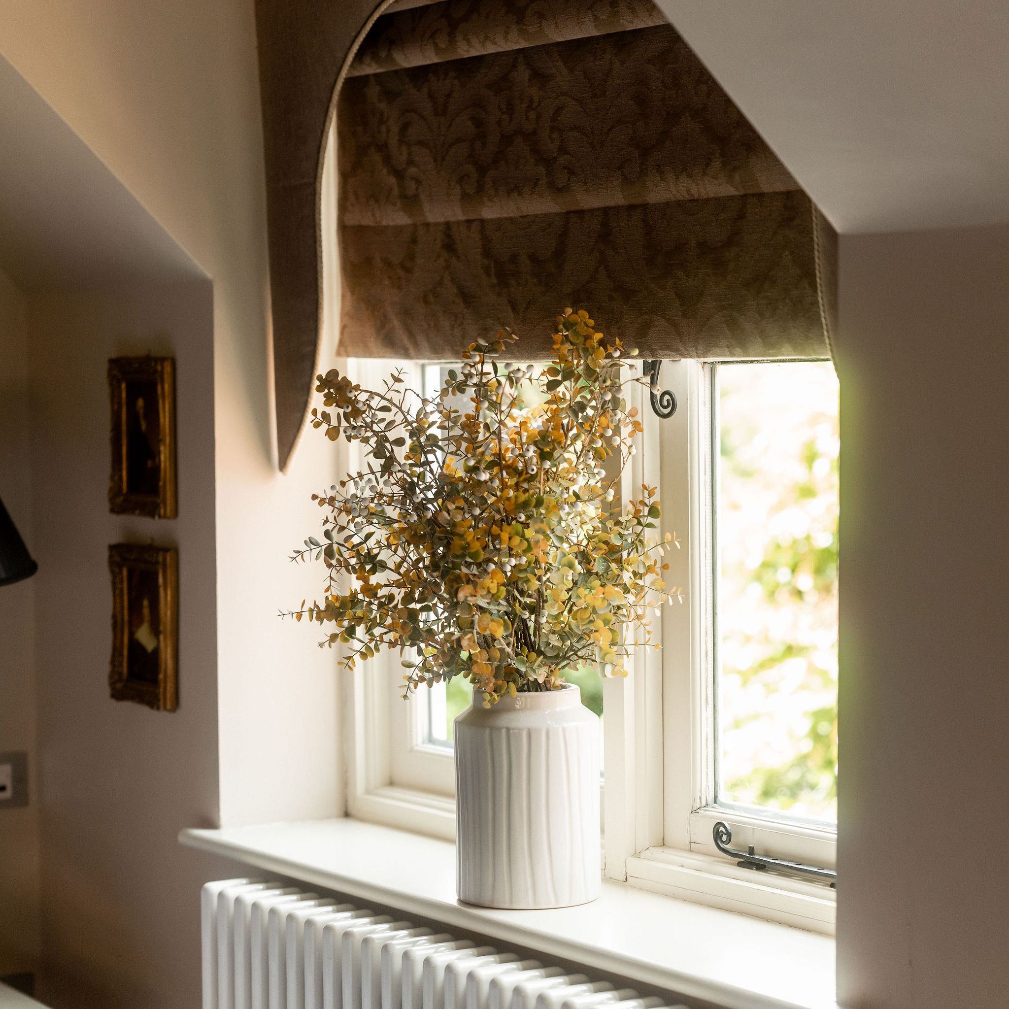 A vase with yellow flowers placed on a windowsill in front of a window with brown roman shades, above a white radiator.