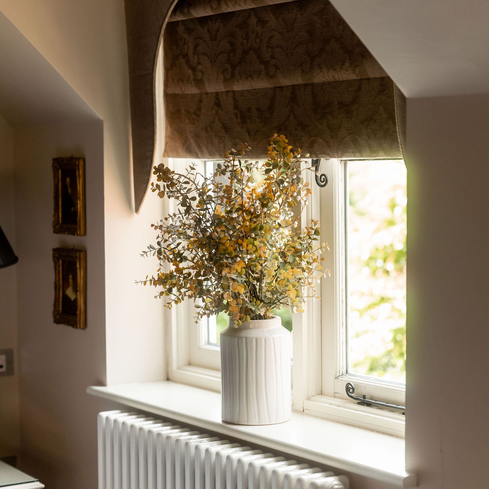 A vase with yellow flowers placed on a windowsill in front of a window with brown roman shades, above a white radiator.