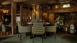 Cozy rustic restaurant interior with wooden beams, brick walls, green upholstered chairs, set dining tables, floral arrangement, and soft lighting.