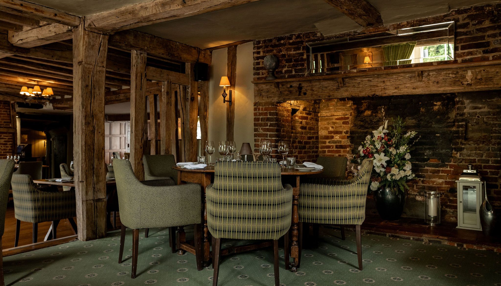 Cozy rustic restaurant interior with wooden beams, brick walls, green upholstered chairs, set dining tables, floral arrangement, and soft lighting.