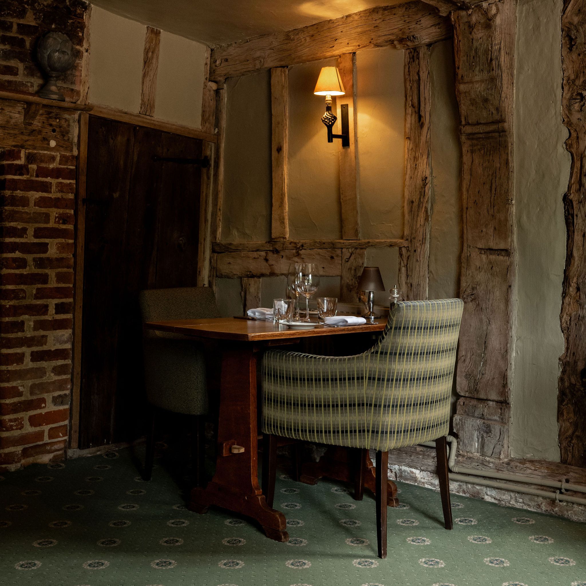 A cozy dining area with a wooden table set for two, upholstered chairs, exposed wooden beams, brickwork, and a wall-mounted lamp in a rustic room.