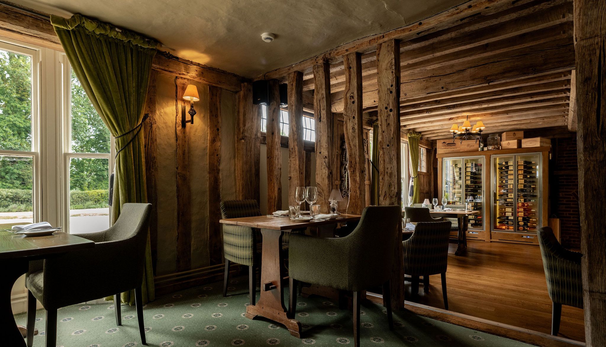 Cozy restaurant interior with rustic wooden beams, green carpet, elegant chairs, set tables, large windows with green curtains, and a well-lit wine display cabinet.