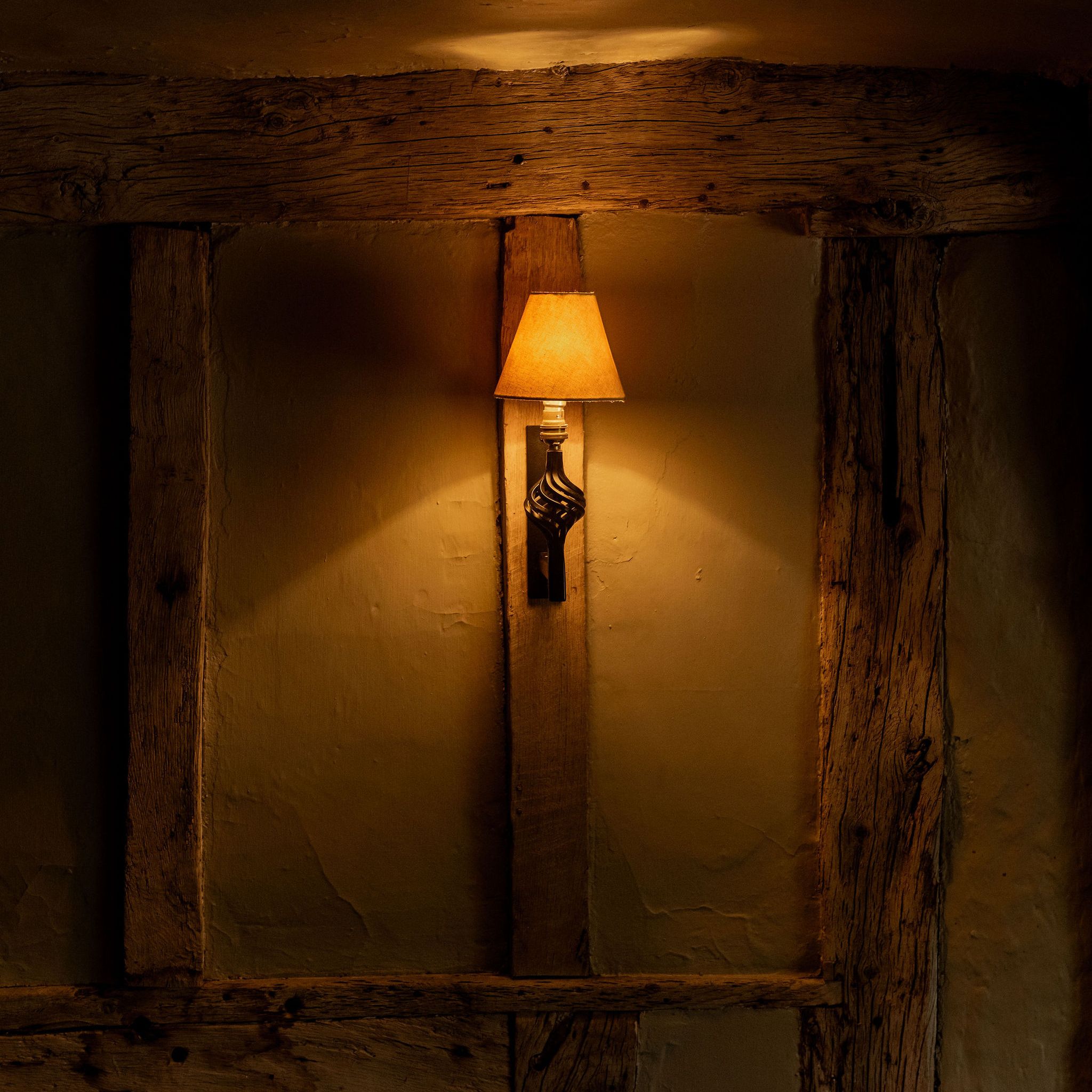 A warmly lit wall sconce with a yellow lampshade mounted on a rustic, wooden beam wall in a dimly lit room.