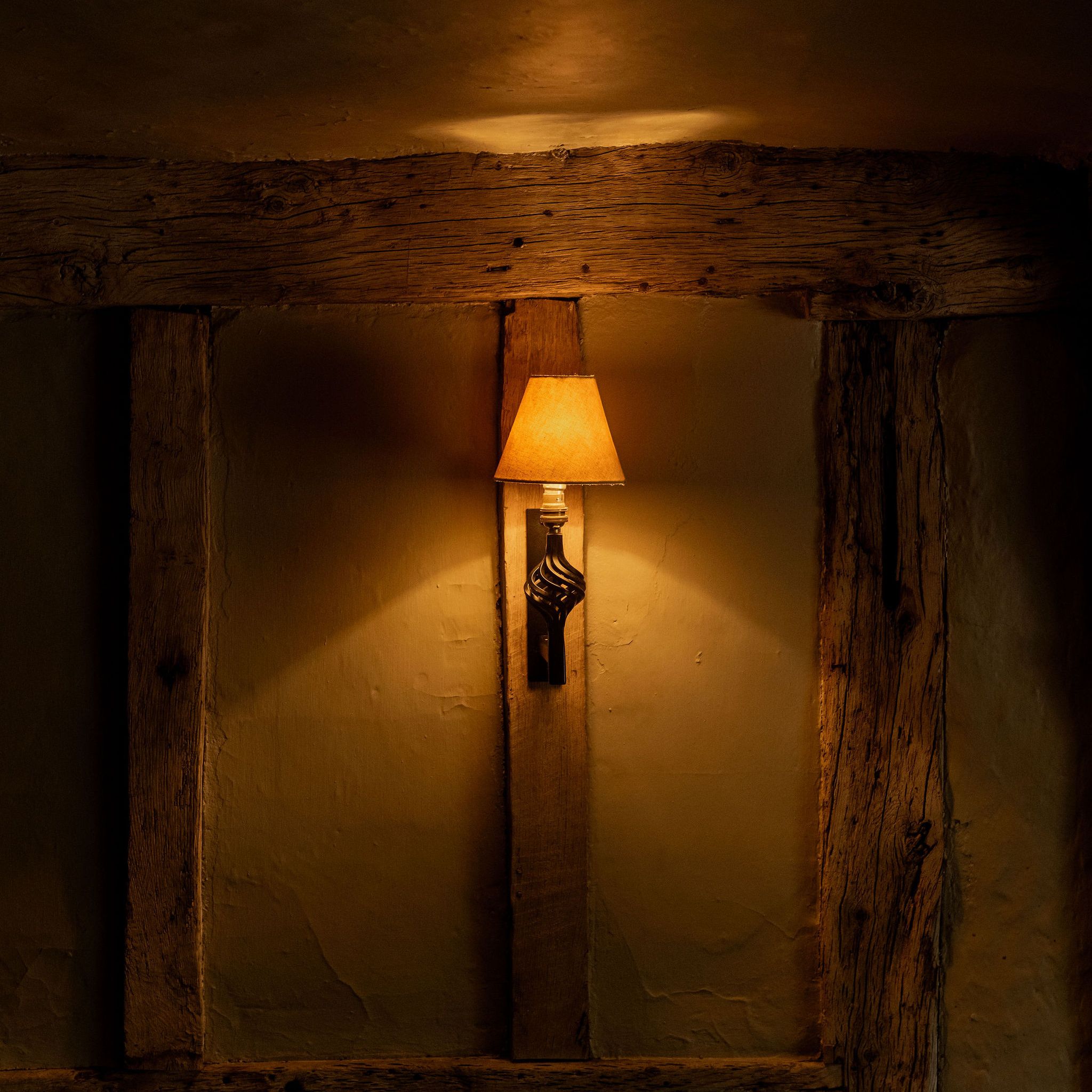 A warmly lit wall sconce with a yellow lampshade mounted on a rustic, wooden beam wall in a dimly lit room.