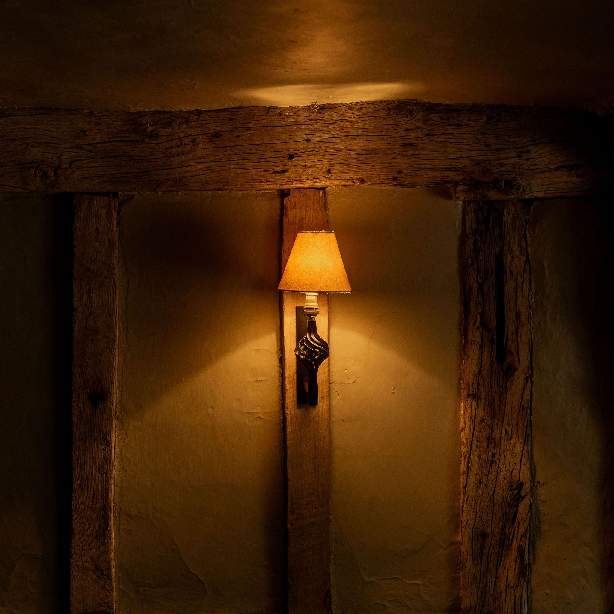 A warmly lit wall sconce with a yellow lampshade mounted on a rustic, wooden beam wall in a dimly lit room.