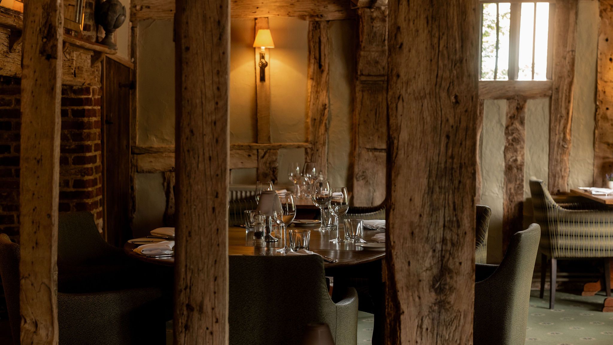 A cozy rustic restaurant interior with exposed wooden beams, brick walls, soft lighting from a wall lamp, and tables set with wine glasses and cutlery.