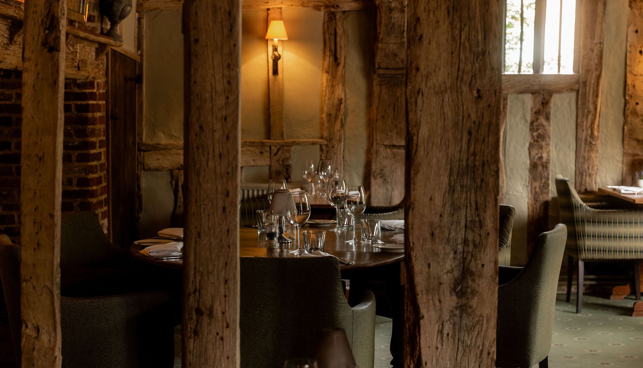 A cozy rustic restaurant interior with exposed wooden beams, brick walls, soft lighting from a wall lamp, and tables set with wine glasses and cutlery.