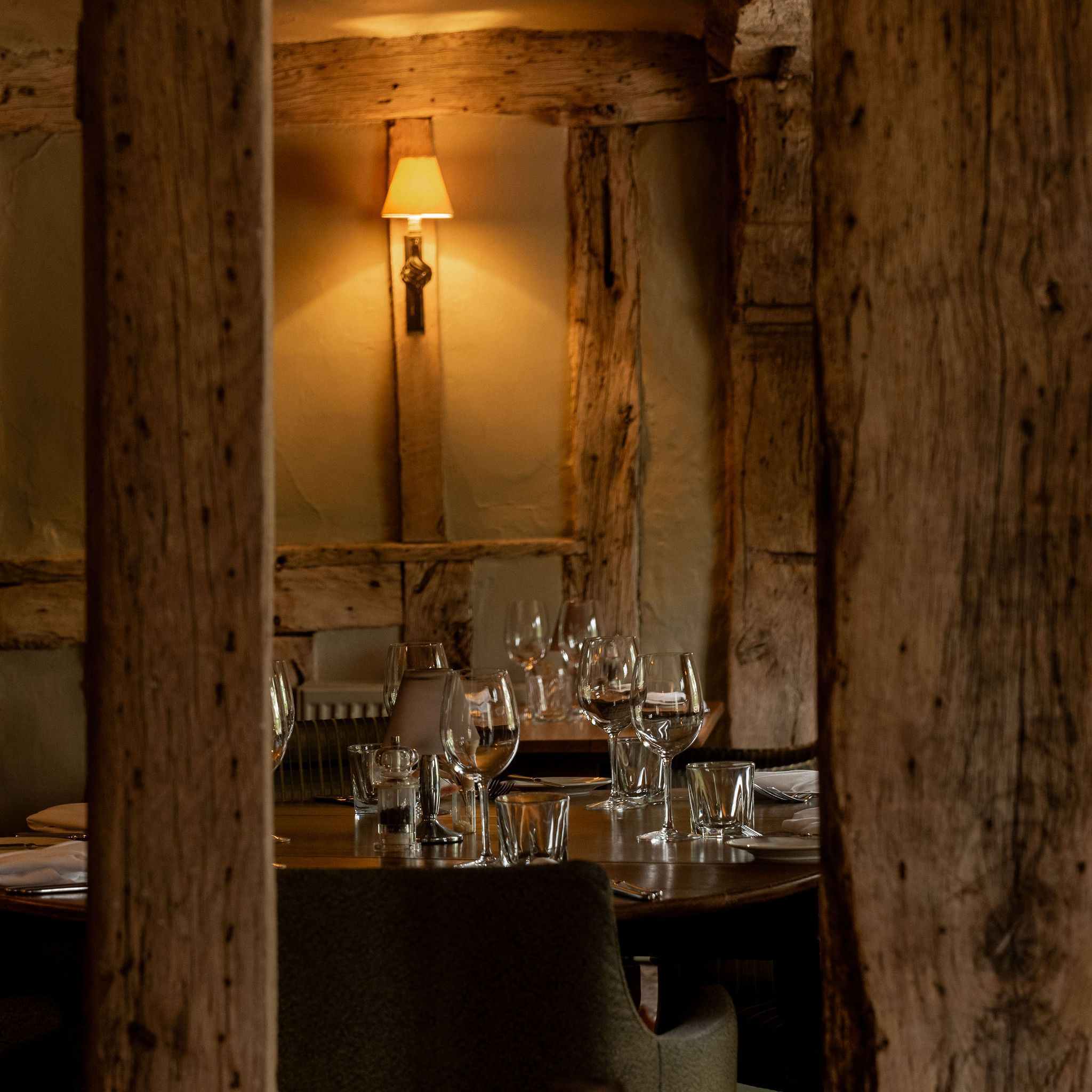 A cozy restaurant interior with rustic wooden beams, a lit wall lamp, and a round table set with empty wine glasses, water glasses, plates, and napkins.