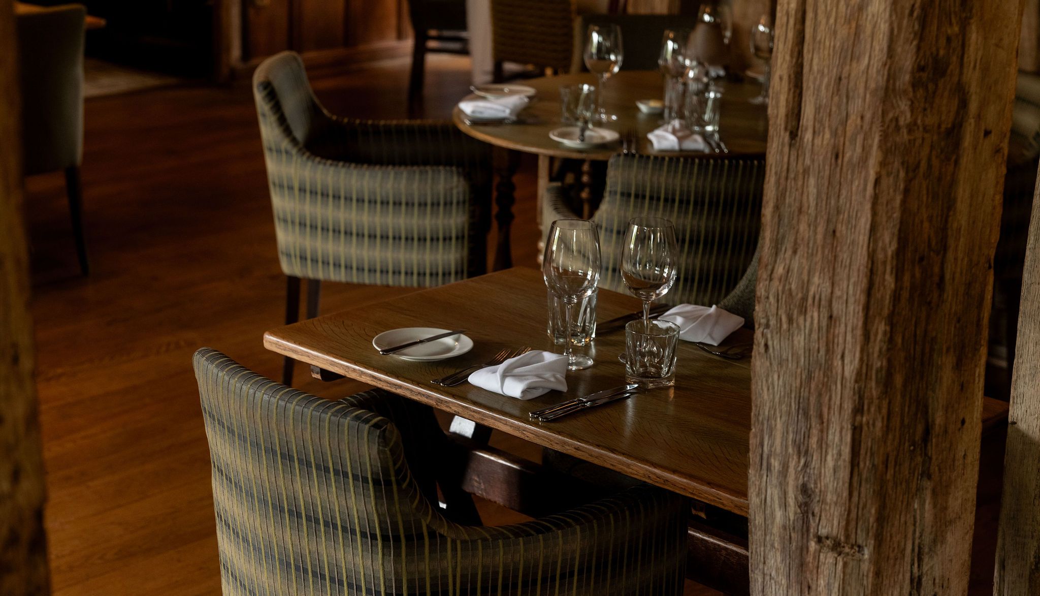 A cozy restaurant setting with wooden tables set for two, featuring striped cushioned chairs, glassware, napkins, and cutlery, on a wood floor with rustic wooden beams in the foreground.