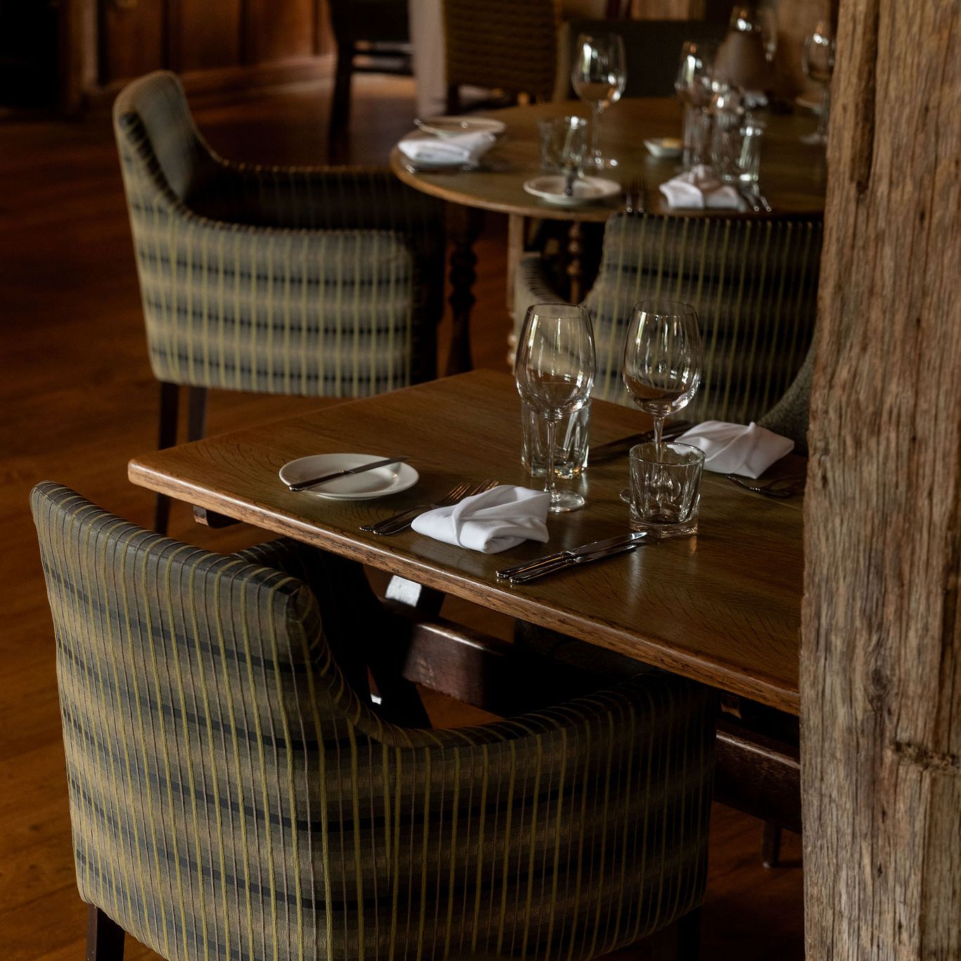 A cozy restaurant setting with wooden tables set for two, featuring striped cushioned chairs, glassware, napkins, and cutlery, on a wood floor with rustic wooden beams in the foreground.